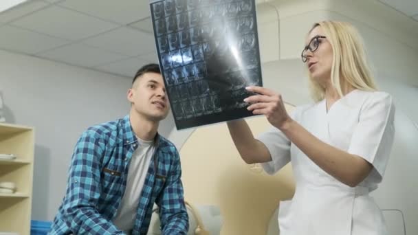 Vrouwelijke arts radioloog legt de resultaten van MRI-scan voor jonge mannelijke patiënt uit, toont de momentopname met beelden, observeert en analyseert MRI-scan in moderne kliniek naast moderne MRI-scanner - Video