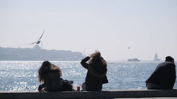 04.03.2021. istanbul. Turkey. Bosphorus istanbul. People and tourist are sitting on concrete stone in shore of bosporus. They are sitting and taking photo and feeding seagulls and doves and pigeons - Photo, Image