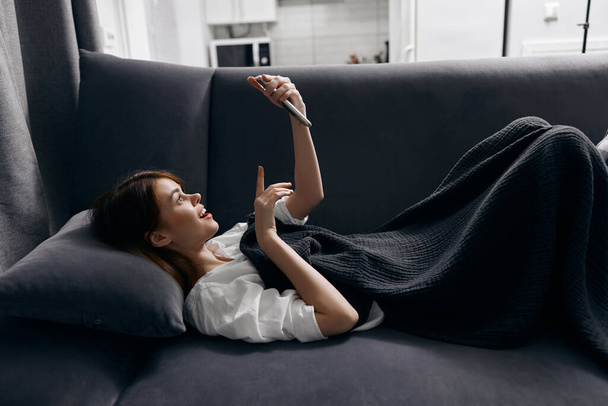 woman looking at the phone screen hiding with a blanket on the sofa - Фото, изображение