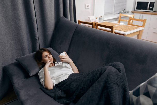 mujer en una camiseta blanca con un teléfono en la mano se encuentra en el sofá interior habitación cómoda - Foto, Imagen