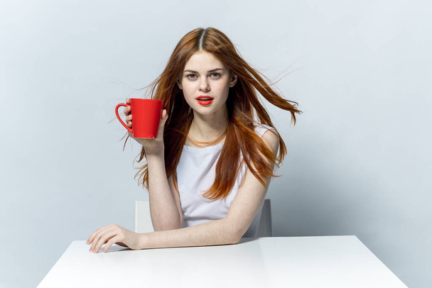 mujer sosteniendo una taza roja en su mano mientras está sentada a la mesa de descanso - Foto, Imagen