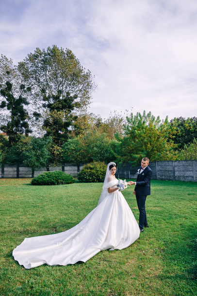Молодий стильний наречений у блакитному костюмі і красива наречена в білій сукні ходять, тримаючи руки в саду з зеленими рослинами на природі. Весільний портрет молодят
. - Фото, зображення