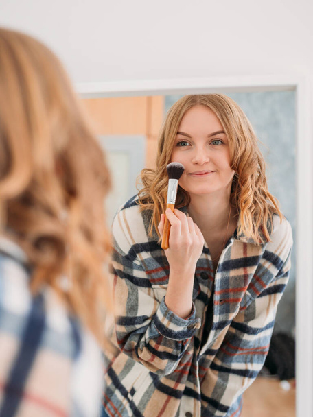 Roztomilá běloška používající štětec na líčení a usmívající se před zrcadlem. Krása, make-up, kosmetika, ranní a lidová koncepce - Fotografie, Obrázek
