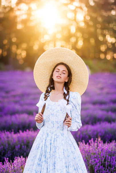 Beautiful girl in a big hat and a full dress on a background of lavender. A young model with braids in her hair. Summer photo in the sun - Photo, Image