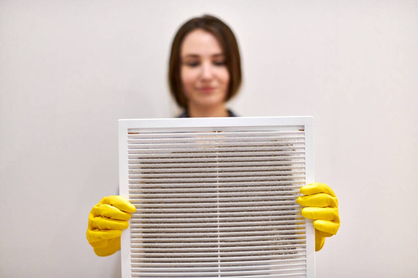 Woman holds ventilation grill with dust filter to clean it. Extremely dirty and dusty white plastic, harmful for health. Housewife in protective yellow rubber gloves and blue-grey uniform, blurred. - Photo, Image