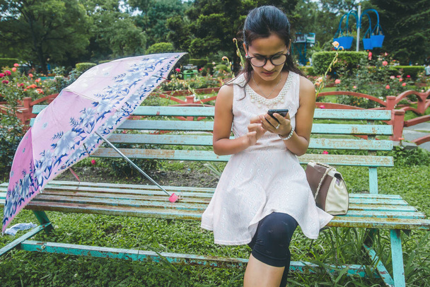 若いですアジアのネパール人の女の子座っていますベンチに傘と使用彼女の電話で公園で待っている間彼女の友人. - 写真・画像