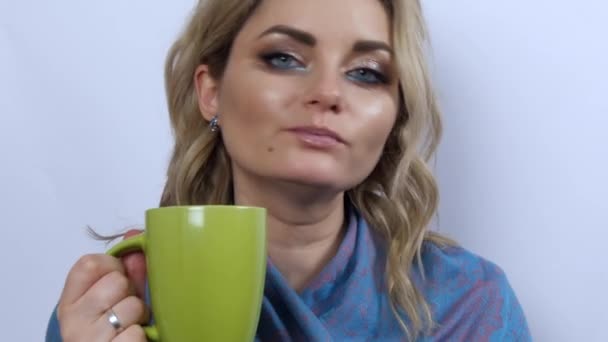 Gyönyörű fiatal nő modell tartja zöld csésze tea vagy kávé a kezében, megissza, élvezi, és úgy néz ki, nyugdíjas fehér háttérrel. Fiatal lány gyönyörű füstös szemfestékkel és kék sállal a vállán - Felvétel, videó
