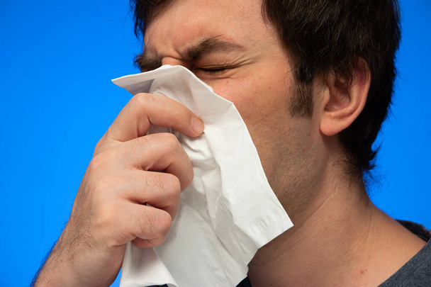 Caucasian male sneezing blowing nose into white paper tissue close up studio shot isolated on blue background. - Photo, Image
