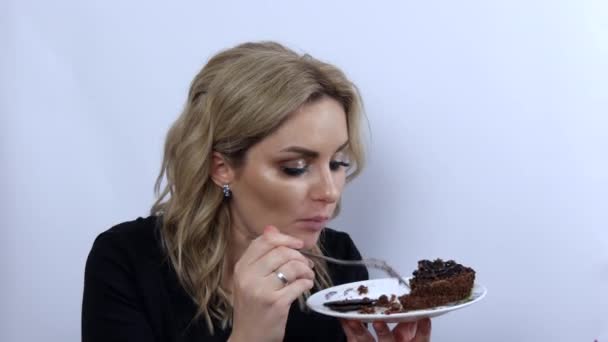 Belle jeune femme modèle drôle manger brownie gâteau au chocolat sur fond blanc. Jeune fille avec beau maquillage des yeux fumé et robe de soirée noire. Vidéo de vitesse - Séquence, vidéo