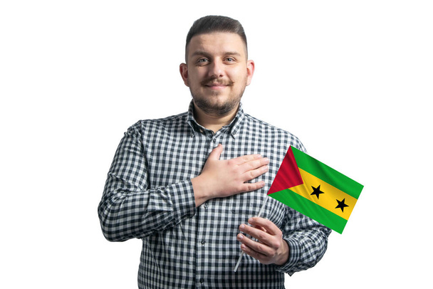 Hombre blanco sosteniendo una bandera de Santo Tomé y Príncipe y sostiene su mano sobre su corazón aislado sobre un fondo blanco. - Foto, imagen