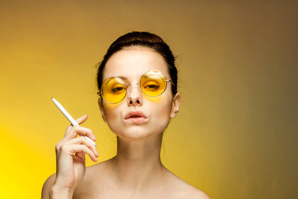 μελαχρινή σε κίτρινα γυαλιά γυμνή ώμους τσιγάρο στα χέρια συναισθήματα κίτρινο φόντο - Φωτογραφία, εικόνα