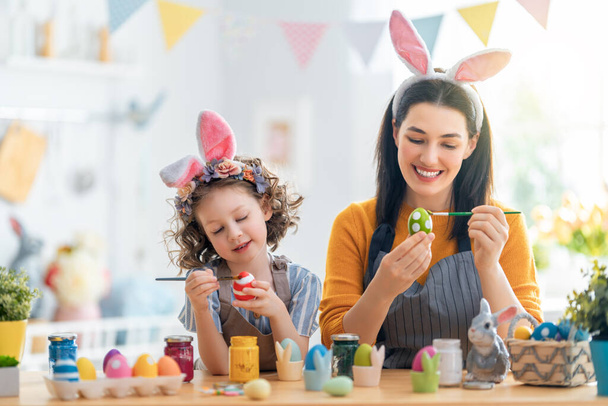 Mother and her daughter painting eggs. Happy family preparing for Easter. Cute little child girl wearing bunny ears. - Foto, immagini