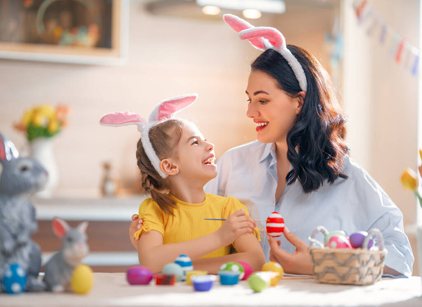 Mother and her daughter painting eggs. Happy family preparing for Easter. Cute little child girl wearing bunny ears. - Valokuva, kuva