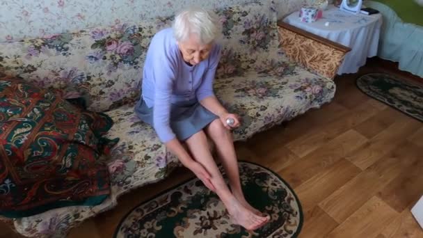 Ηλικιωμένη γυναίκα που βάζει κρέμα στα πόδια της καθισμένη στον καναπέ - Πλάνα, βίντεο