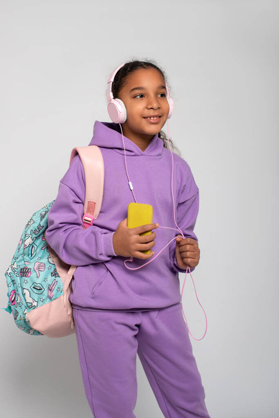 Porträt eines jungen dunkelhäutigen afrikanischen Mädchens mit fliederfarbenem Trainingsanzug im Stehen oder Gehen mit rosa Kopfhörern und gelbem Telefon. Lebensstil von Kindern und Erwachsenen. - Foto, Bild