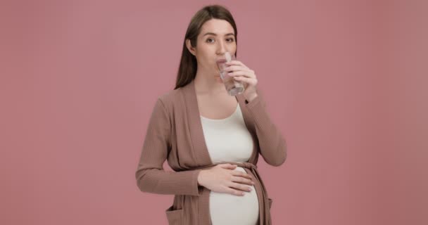 Водный баланс для хорошей беременности. Веселая беременная женщина пьет стакан воды, трогает живот, розовый фон - Кадры, видео
