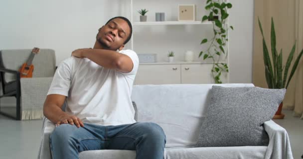Afro-americano homem jovem etnia cara em roupas casuais senta-se no sofá em casa em posição desconfortável sente desconforto espasmo dor no pescoço sofre de doença crônica massageando os músculos das costas com a mão  - Filmagem, Vídeo