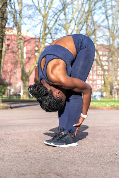 Black African woman warming up and doing back and leg stretches bending over holding her legs to start doing sports in a public park on a sunny day - Photo, Image