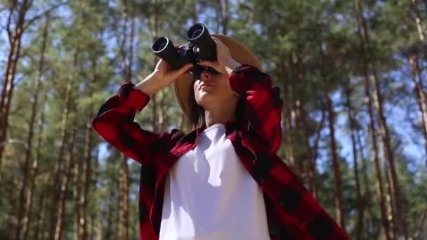 Una chica con sombrero mira a través de los prismáticos en el bosque. Concepto caminata, turismo. - Metraje, vídeo