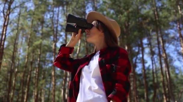 Ένα κορίτσι με καπέλο κοιτάει μέσα από κιάλια στο δάσος. Concept πεζοπορία, τουρισμός. - Πλάνα, βίντεο
