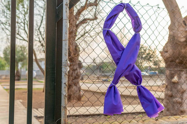 Símbolo de una cinta púrpura hecha de tela y atada con bridas a una puerta de metal de un centro educativo cerrado y desenfocado. Símbolo conmemorativo del Día Internacional de la Mujer, 8 de marzo - Foto, Imagen