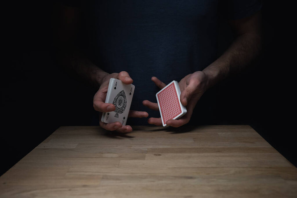 Muž v modré košili na tmavém pozadí, držící rozštěpený balíček červených karet u dřevěného stolu s pikovým esem. Games cards playing poker gambling. - Fotografie, Obrázek