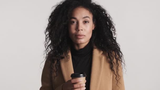Photo studio d'une jolie femme afro-américaine aux cheveux bruns tenant une tasse de café chaud regardant intensément dans une caméra isolée sur fond blanc. Café à emporter concept - Séquence, vidéo