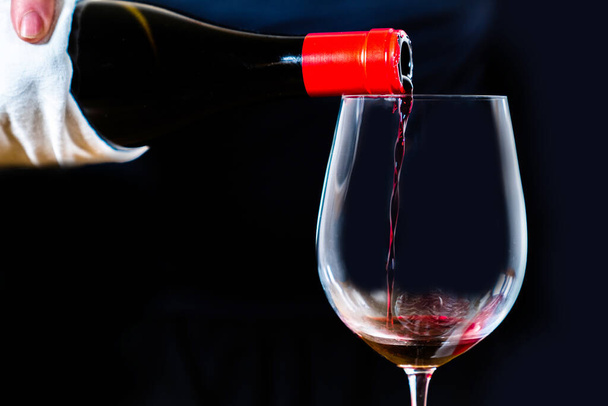 Névtelen sommelier spanyol fekete bort szolgál fel elegáns kristálypohárban, fekete háttérrel. Piros bor öntése palackból a borosüvegbe fekete alapon. - Fotó, kép