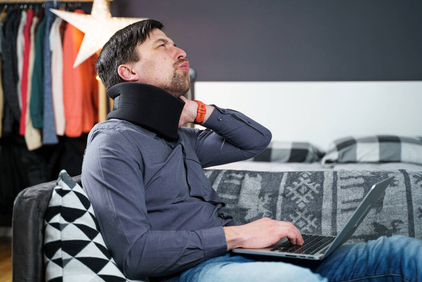 Man employee after accident working at home behind laptop and feels pain in neck, uses black neck collar support. Male with neck injury freelance programmer on sick leave working at computer on couch. - Photo, Image