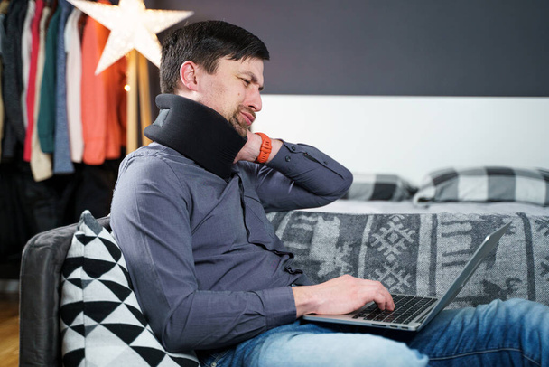 Κουρασμένος ταλαιπωρημένος άνθρωπος που υποφέρει από πόνο στον αυχένα ενώ εργάζεται στον υπολογιστή. Αντρικό μασάζ στο λαιμό, φοράει κολάρο λαιμού, κάθεται στον καναπέ, υποφέρει από δυσφορία, πολλές ώρες καθιστικής υπερεργασίας - Φωτογραφία, εικόνα