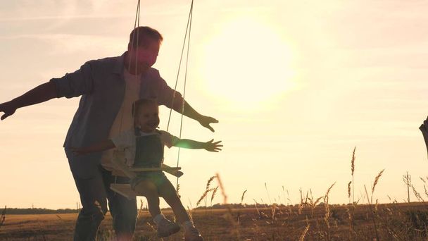 Joyeux petit enfant avec un ours en peluche et papa monte une balançoire au coucher du soleil dans le parc, jouant l'avion volant dans les airs. Fille et père s'amusent à l'aube. Joyeux concept de vie familiale. Enfant. - Photo, image