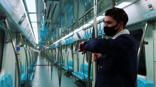 手時計と彼の携帯電話を見て- -黒いマスクの若いハンサムな男が空の地下鉄の列車に立っている - 映像、動画