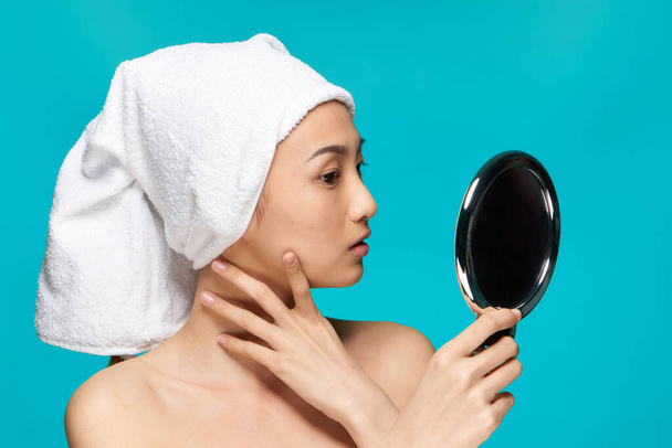 femme d'apparence asiatique avec les épaules nues regarde miroir avec une serviette sur son fond bleu tête - Photo, image