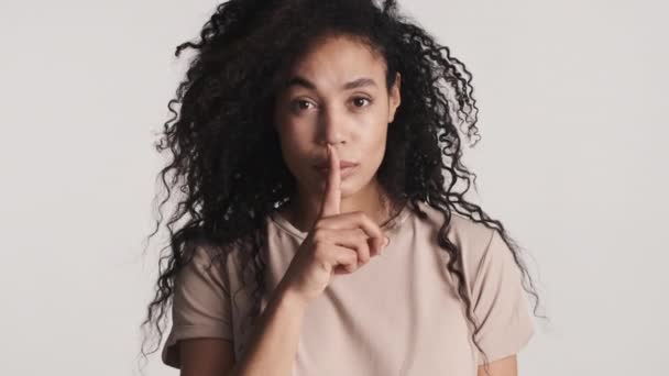 Genç Afro-Amerikalı bir kadın ön parmağını dudaklarının üzerinde tutuyor. Kendine güveniyor gibi görünüyor. Beyaz arka planda gizli bir ifade olduğunu gösteriyor. Söyleme. - Video, Çekim
