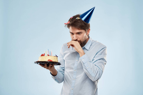Homem barbudo com um prato de bolo em um fundo azul e um chapéu de festa de aniversário na cabeça - Foto, Imagem
