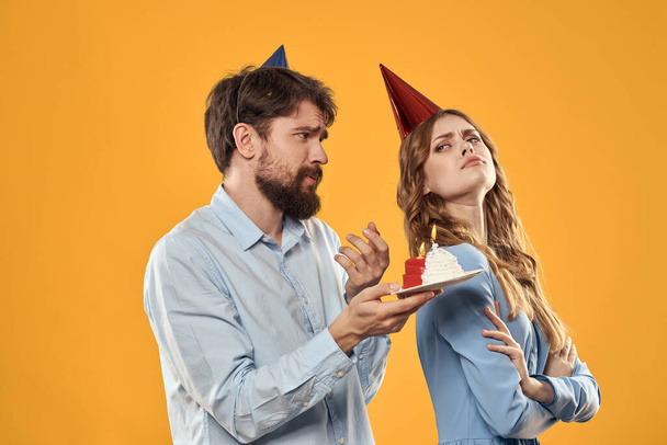 Γεννέθλια κόμμα άνθρωπος και γυναίκα σε ένα καπέλο με ένα κέικ σε κίτρινο φόντο περικοπή άποψη - Φωτογραφία, εικόνα