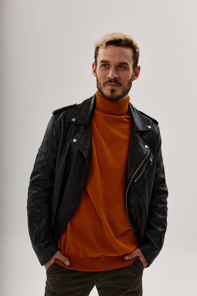 egy férfi narancssárga pulóverben és bőrdzsekiben a zsebében tartja a kezét könnyű háttérrel. - Fotó, kép