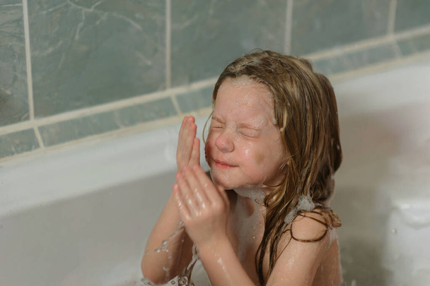 bambina si pulisce il viso dalla schiuma mentre fa il bagno nel bagno - Foto, immagini