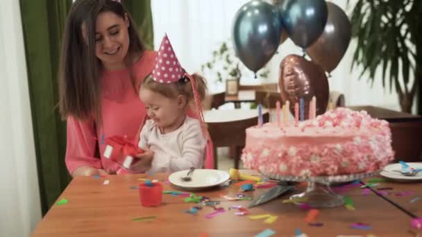 Η μαμά και η κόρη κάθονται στο σπίτι στο τραπέζι κοντά στην τούρτα και γιορτάζουν τα γενέθλιά τους. Το κορίτσι κρατάει ένα κόκκινο κουτί στα χέρια της.. - Πλάνα, βίντεο
