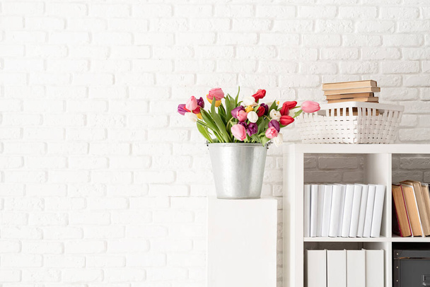 白いレンガの壁の背景に本棚の横に新鮮なチューリップの花のバケツ - 写真・画像