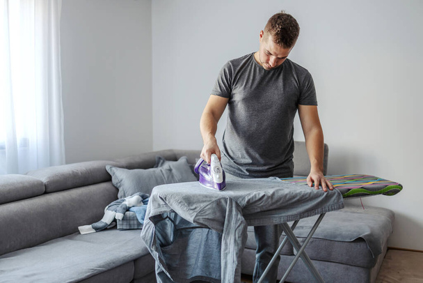 Dělení domácích prací, žehlení. Muž středního věku nespokojen s tím, že musí žehlit šaty. Váhavě žehlí hromadu prádla v obýváku. Svobodný muž nebo dobrý manžel - Fotografie, Obrázek