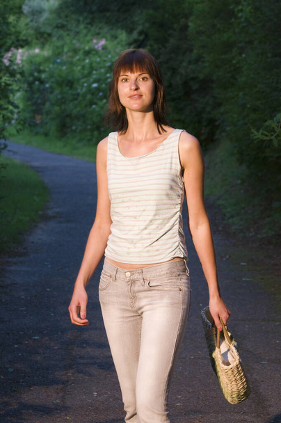 Przedni widok z trzech czwartych młodej, brunetki w dżinsach i koszulce z torbą spacerującą po leśnej ścieżce asfaltowej i wyglądającą przyjaźnie w kamerze.  - Zdjęcie, obraz