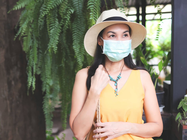 Asiática turista feminina vestindo camisa amarela sem mangas, chapéu e máscara protetora médica, de pé no jardim.. Covid19 prevenção durante a viagem. - Foto, Imagem
