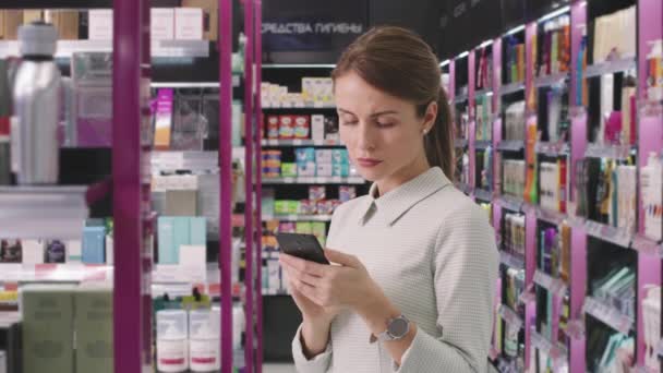 Slow-motion médio tiro de bela mulher caucasiana usando smartphone em pé na loja de cosméticos brilhantes, olhando ao redor ao comprar produtos de beleza - Filmagem, Vídeo
