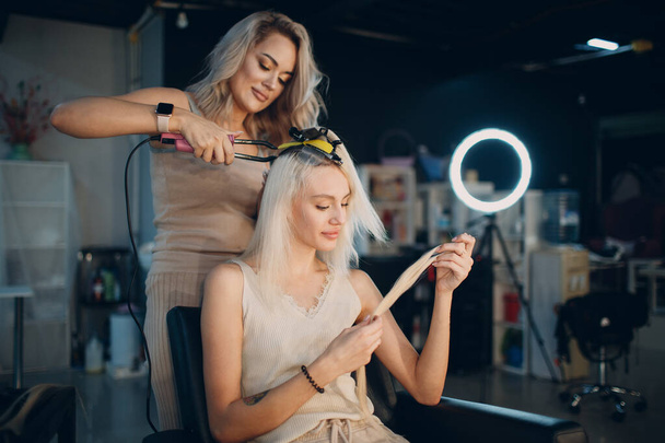 Fryzjer kobieta robi przedłużanie włosów do młodej kobiety z blond włosami w salonie piękności. Profesjonalne przedłużanie włosów. - Zdjęcie, obraz
