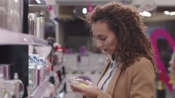 Side-view medium closeup με slowmo της όμορφης γυναίκας με μακριά σγουρά μαλλιά επιλέγοντας νέο άρωμα στο κατάστημα ομορφιάς - Πλάνα, βίντεο