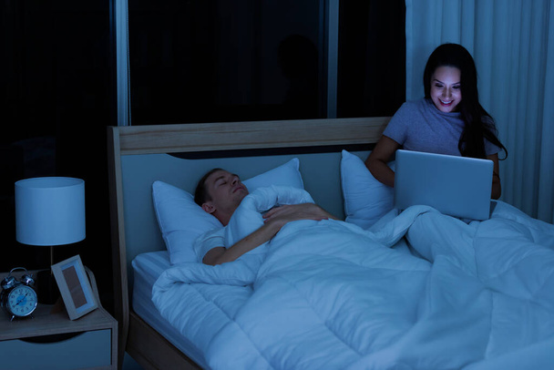 Nainen istuu sängyssä yöllä ja käyttää kannettavaa tietokonetta, kun aviomies nukkuu hänen vieressään. - Valokuva, kuva