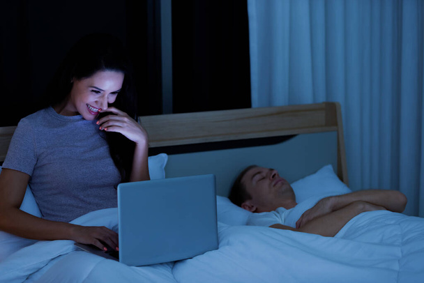 Frau sitzt nachts auf Bett und benutzt Laptop-Computer, während Mann neben ihr schläft. - Foto, Bild