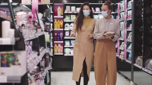 slow-motion medium shot van vrouwelijke schoonheid adviseur en aziatische vrouw in maskers lopen langs planken met schoonheidsproducten in grote cosmetica winkel - Video