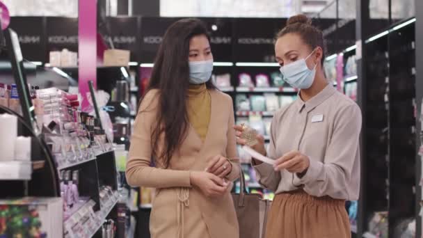 Kozmetik mağazasında yeni parfüm arayan zarif Asyalı bir kadının orta ölçekli çekimi. Kadın güzellik danışmanı test etmek için bir parfüm püskürtüyor. Hepsi maske takıyor. - Video, Çekim